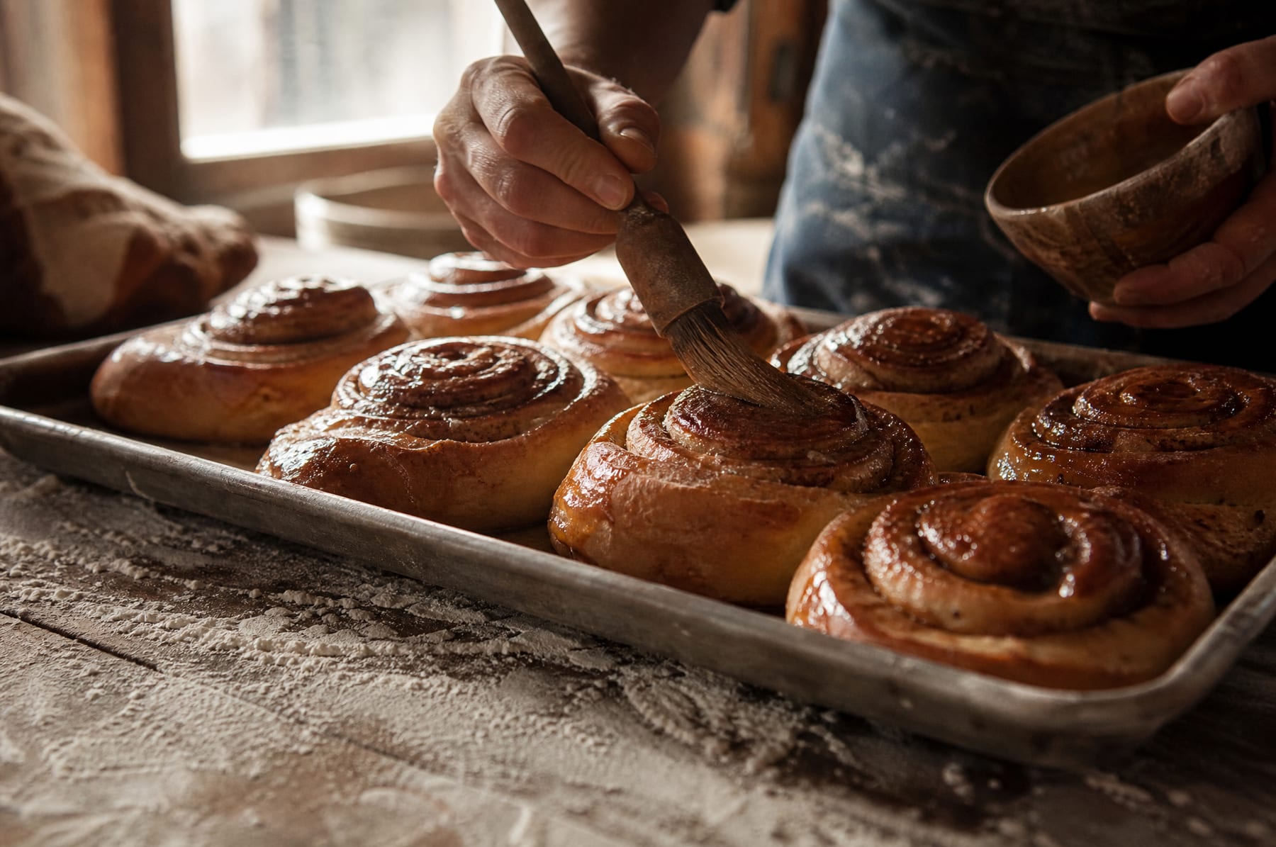 Une plaque de cinnamon rolls dorés à la sortie du four, une main qui les glace au pinceau.